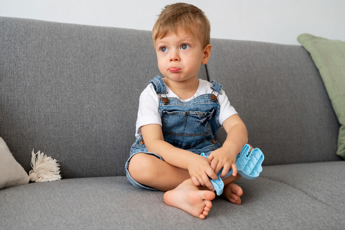 Young kid sitting on a gray couch holding a fidget toy, showing a quirky expression that seems passed down from parents.