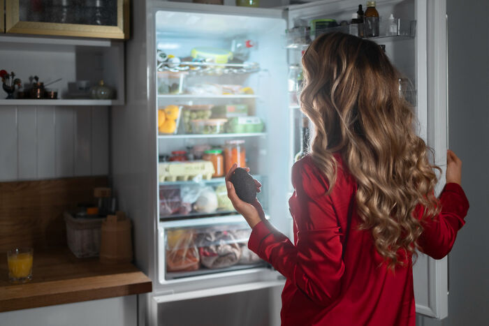 Woman in red pajamas holding an avocado in front of an open fridge, showing kids doing something weird passed down from parents