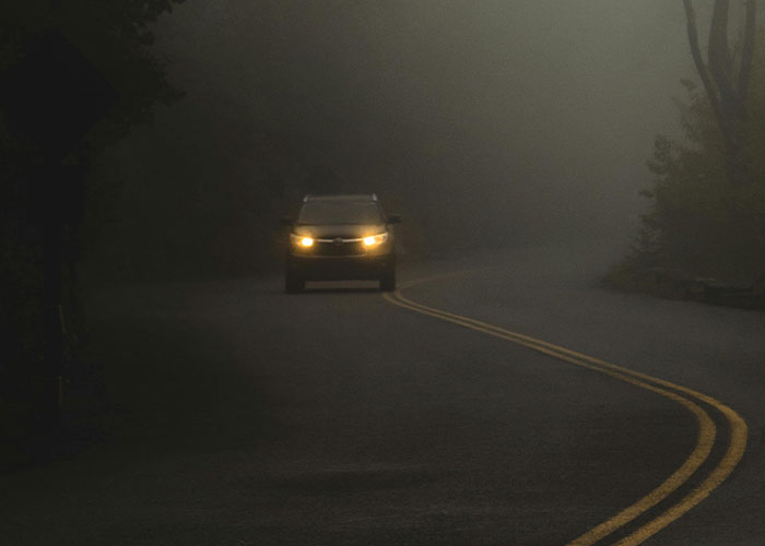 Car driving with headlights on along a foggy winding road, evoking bizarre events that creep people out.