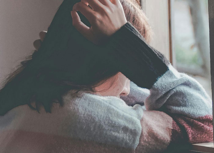 A person covering their face with their hand, looking out a window, evoking feelings from bizarre events stories.