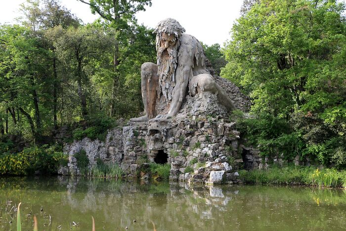 Large stone sculpture of a seated man integrated into natural rocks by a tranquil pond, surrounded by lush green forest.