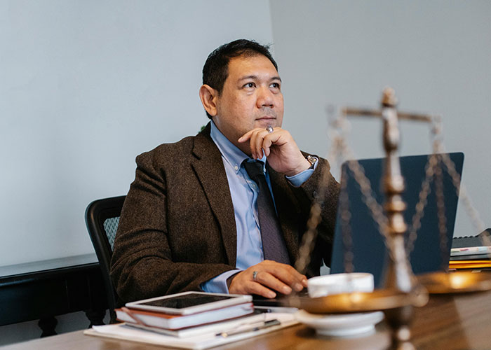 Man in brown blazer at desk, thoughtfully sharing life hacks with laptop and legal scales in an office setting.