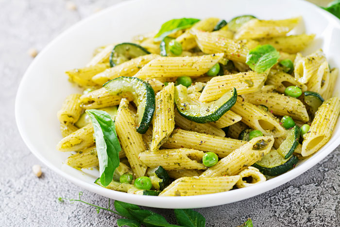 Penne pasta with pesto sauce, zucchini slices, and green peas served in a white bowl on a gray surface. Penne pasta with pesto sauce, zucchini slices, and green peas served in a white bowl on a gray surface.