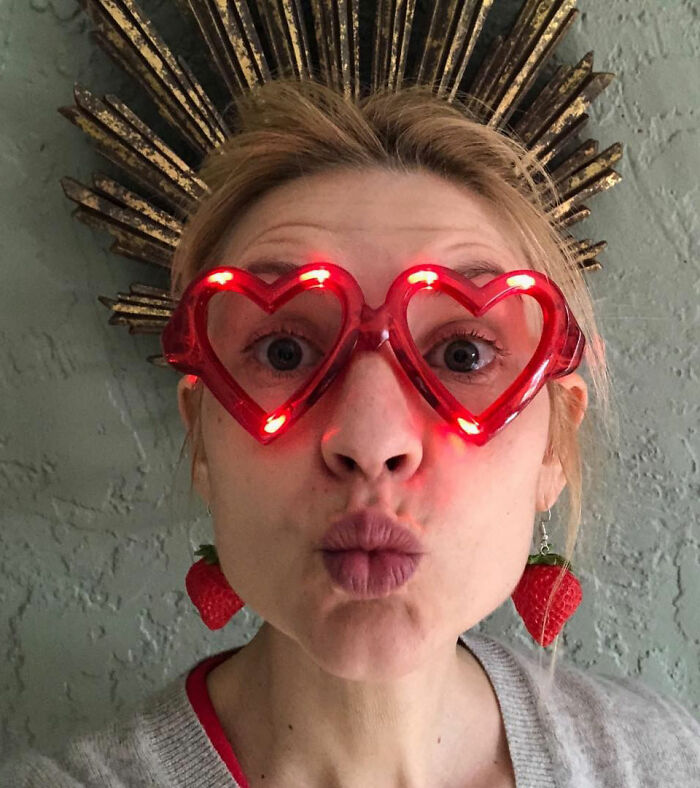 Woman with quivering chin wearing red heart-shaped light-up glasses and strawberry earrings in a playful close-up portrait.
