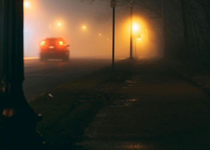 Car driving down a foggy, dark street illuminated by eerie yellow streetlights creating a bizarre and creepy atmosphere.