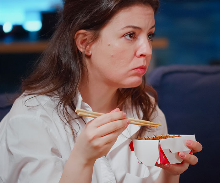 Woman eating leftovers with chopsticks, looking pensive indoors, reflecting themes of weight gain and relationship tension.