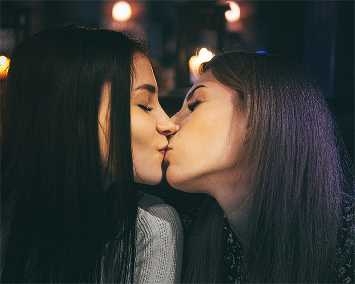 Two women sharing a kiss in a dimly lit setting, highlighting a girlfriend kisses another woman moment.