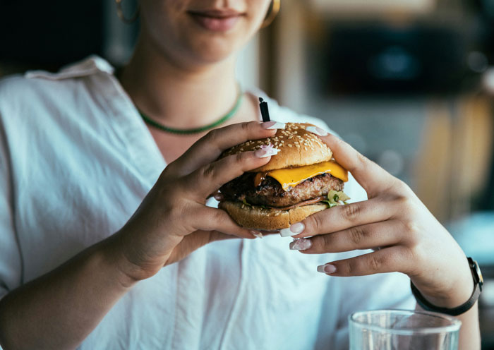 Woman eating a cheeseburger she shouldn&rsquo;t have, coworker paying the price in a casual dining setting.