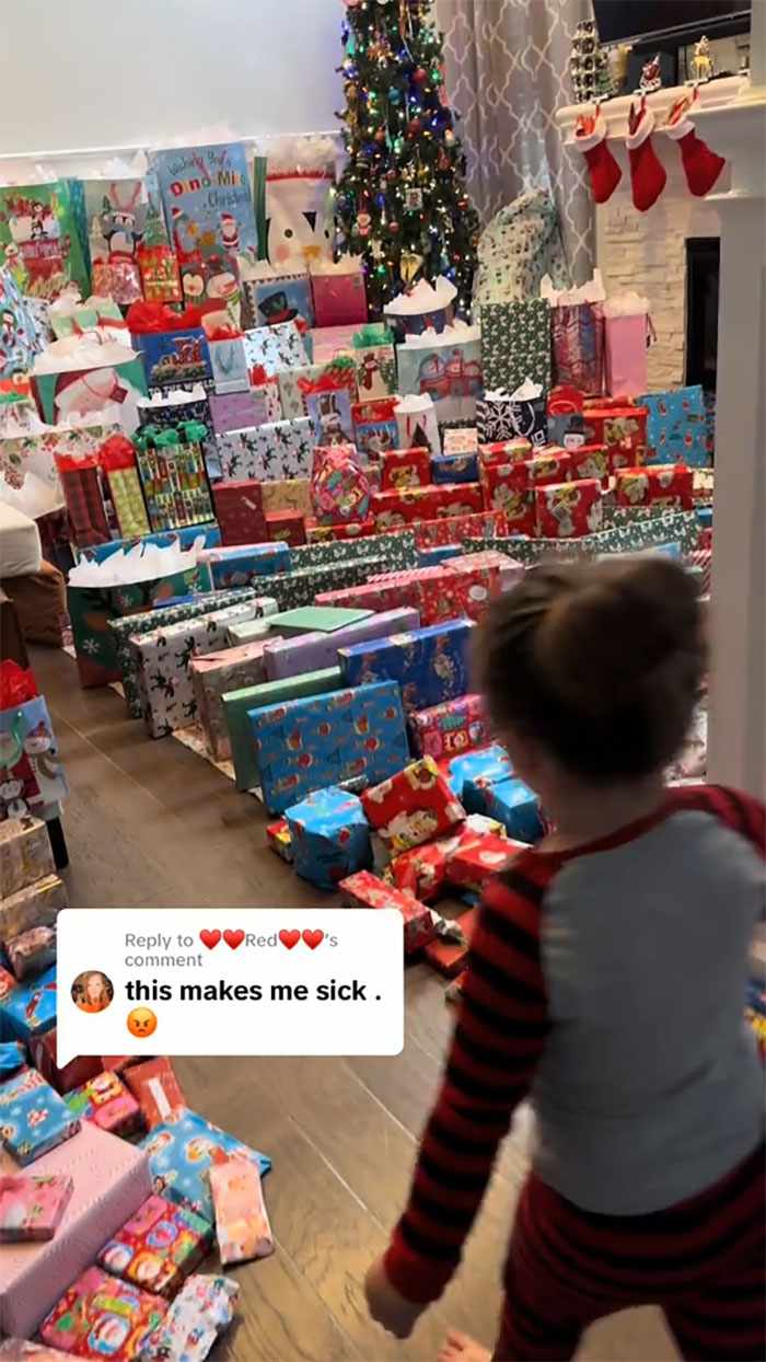Child in pajamas looking at an insane amount of gifts stacked by a Christmas tree, highlighting consumerism backlash for kids.
