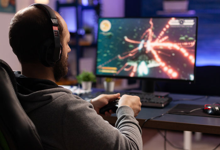 Man wearing headphones playing video games on computer, avoiding Thanksgiving shopping pressure from wife&rsquo;s ban decision.