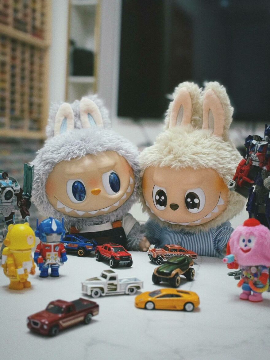 Two original Labubu dolls with large eyes and bunny ears are surrounded by toy cars and action figures on a table.