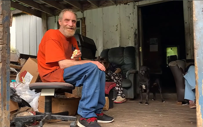 Man in an orange shirt sitting on a chair in a cluttered room with dog and people in the background, viral fame reaction scene