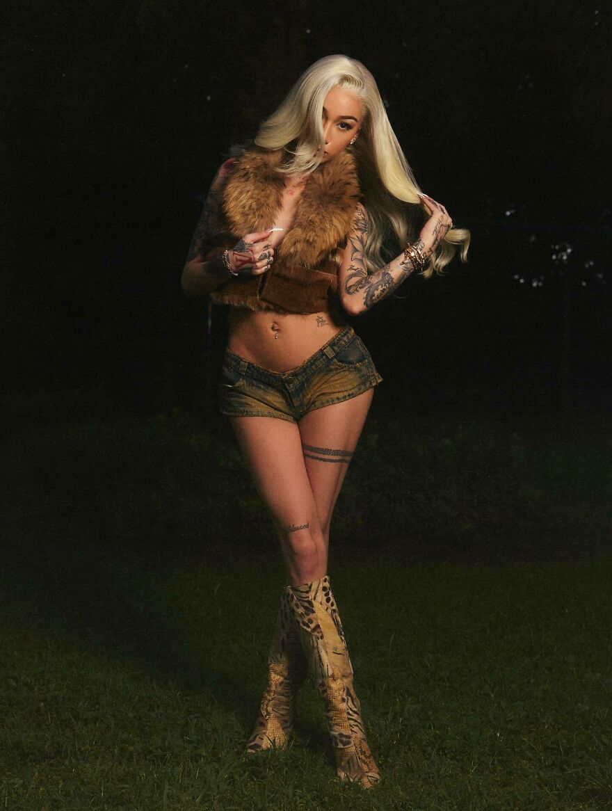 Bhad Bhabie posing outdoors at night, wearing a fur vest, denim shorts, and patterned boots showing a confident style.