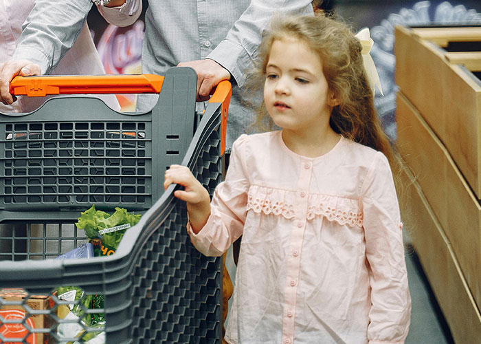 A young girl with long hair in a pink dress standing beside a shopping cart during a grocery store visit.