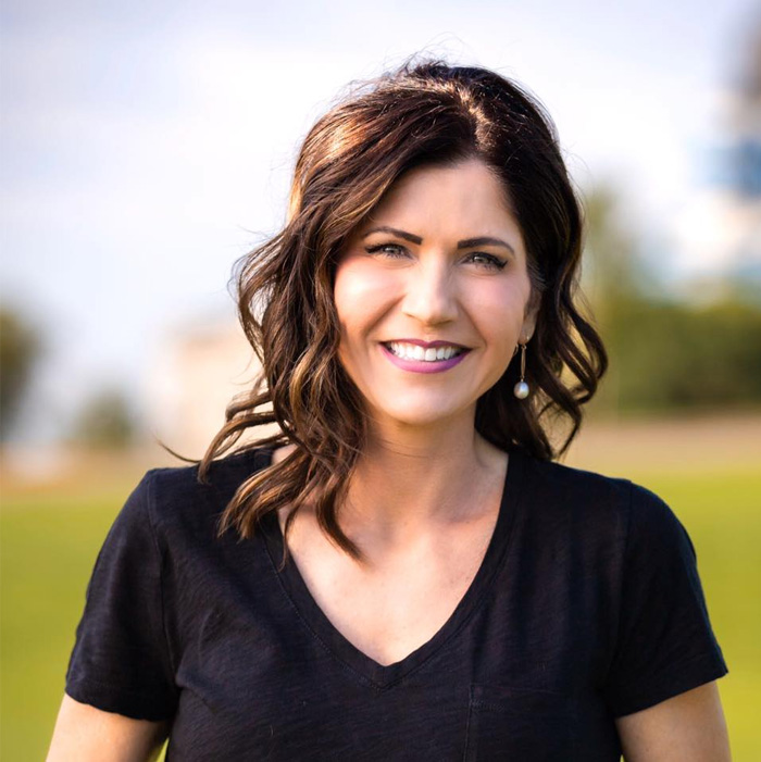 Kristi Noem smiling outdoors with wavy hair, wearing a black shirt and pearl earrings in a natural setting. Kristi Noem smiling outdoors with wavy hair, wearing a black shirt and pearl earrings in a natural setting.