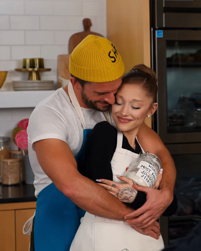 Ariana Grande and Jonathan Bailey hugging in a kitchen, sharing a warm and intimate moment while baking together.