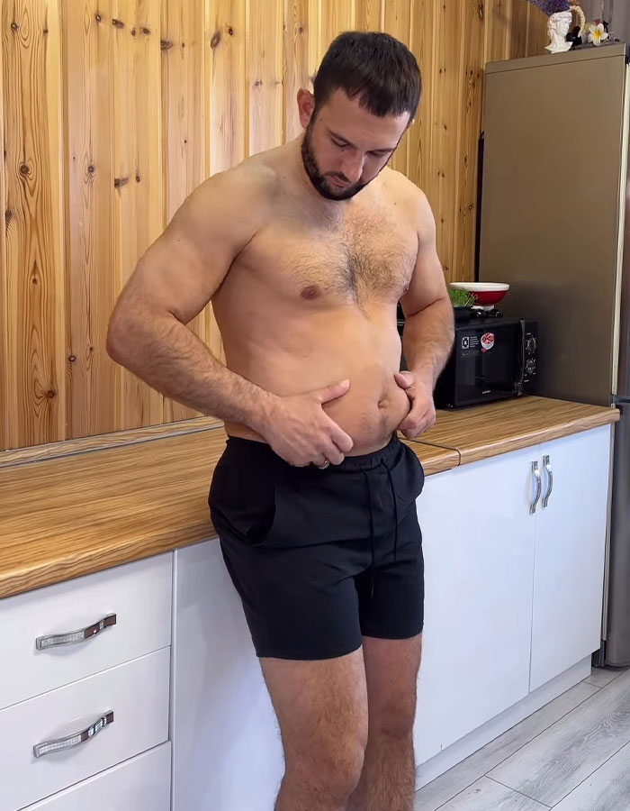 Fitness coach shirtless in kitchen, examining his stomach while wearing black shorts, wooden panel background.