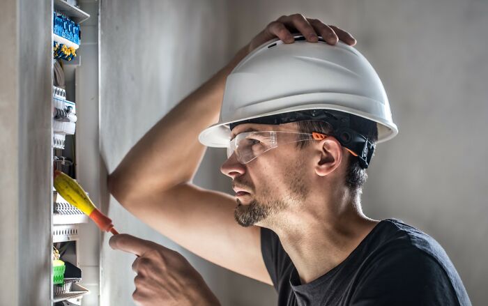 Man wearing safety glasses and a hard hat, examining an electrical panel, illustrating little lie consequences concept.