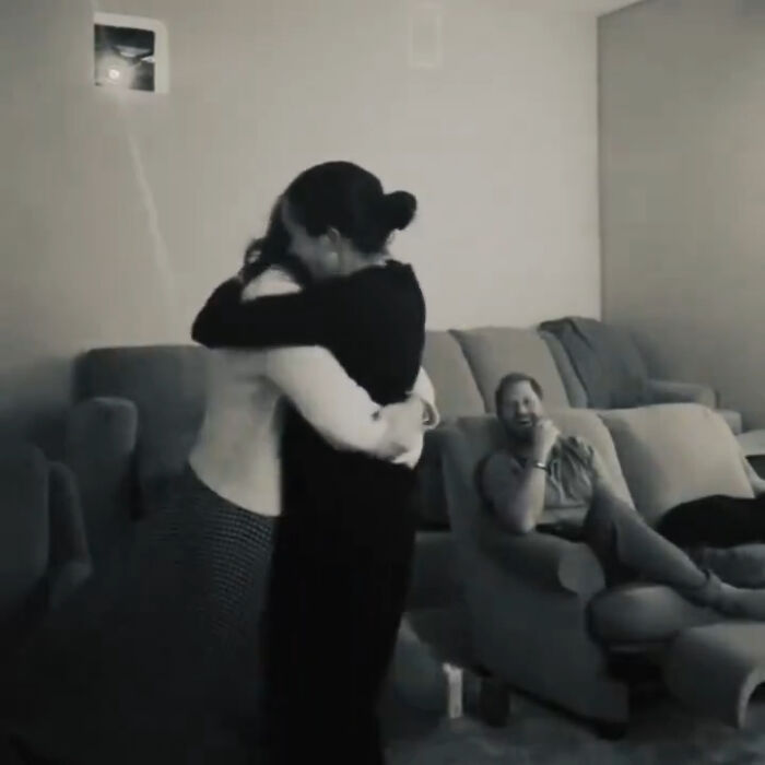Meghan Markle and Prince Harry sharing a hug during a home celebration, with Prince Harry watching and smiling on the couch.