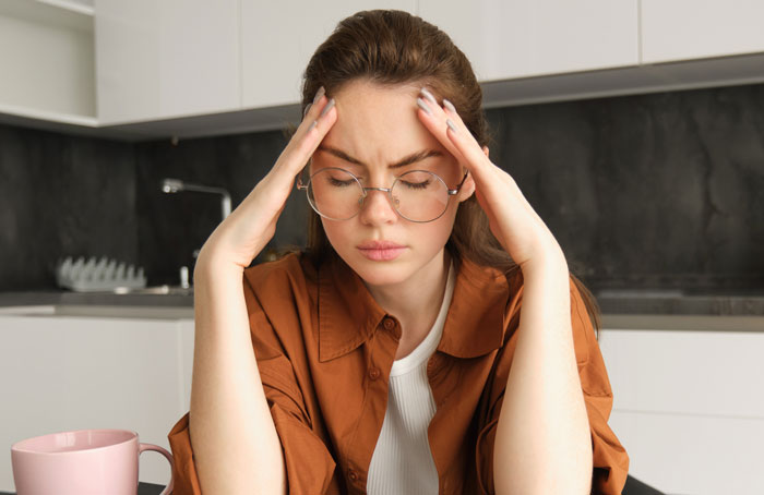 Stressed woman wearing glasses in kitchen holding her head, reflecting Thanksgiving shopping pressure and family tension.