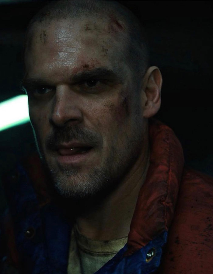 David Harbour with facial bruises wearing a red and blue jacket in a dark scene from Stranger Things set.