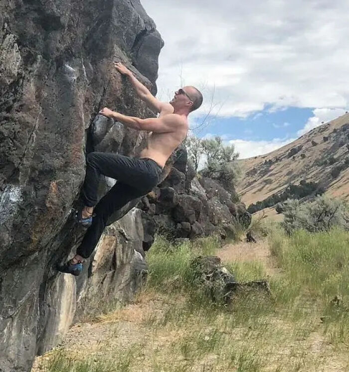 Man climbing a rocky cliff outdoors with mountains and grassland in the background, related to father and son zip-lining incident.