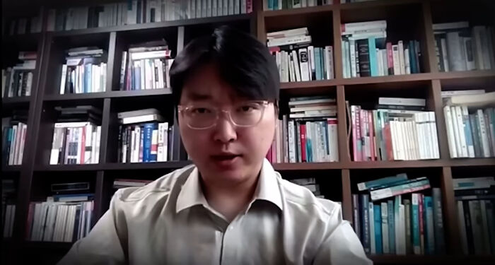 Man wearing glasses and white shirt speaking in front of a bookshelf, related to South Korea asylum and smartest man topic.