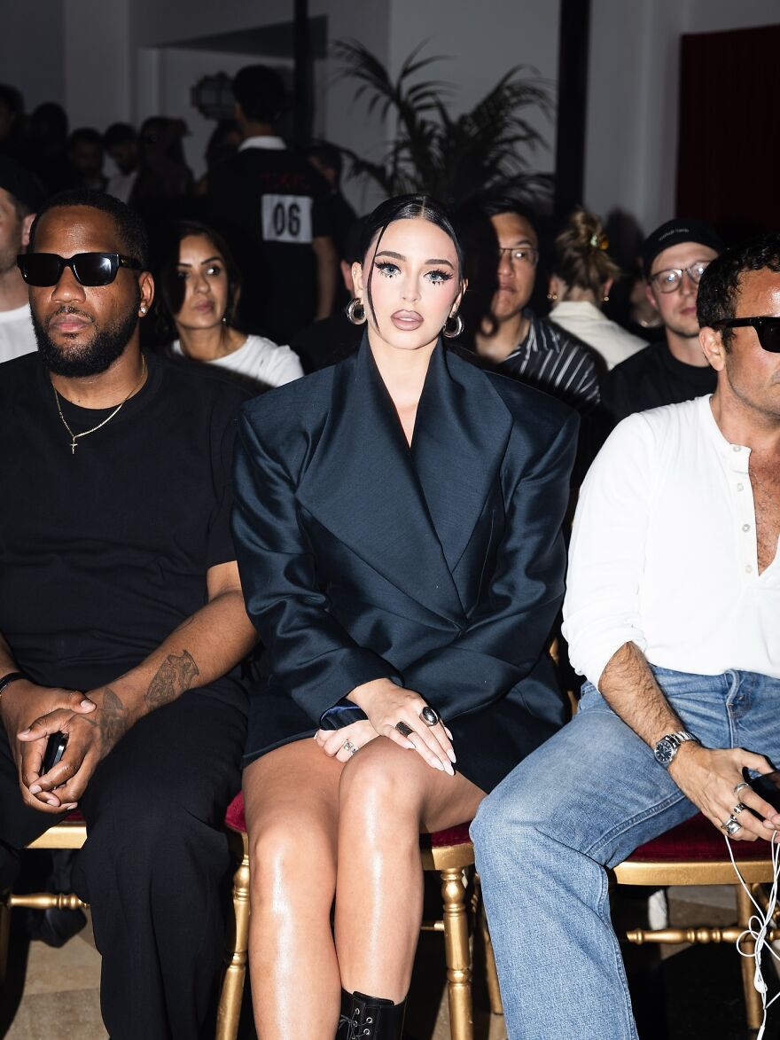 Camilla Araujo sitting front row at an event, wearing a black oversized blazer, showcasing her net worth and style.