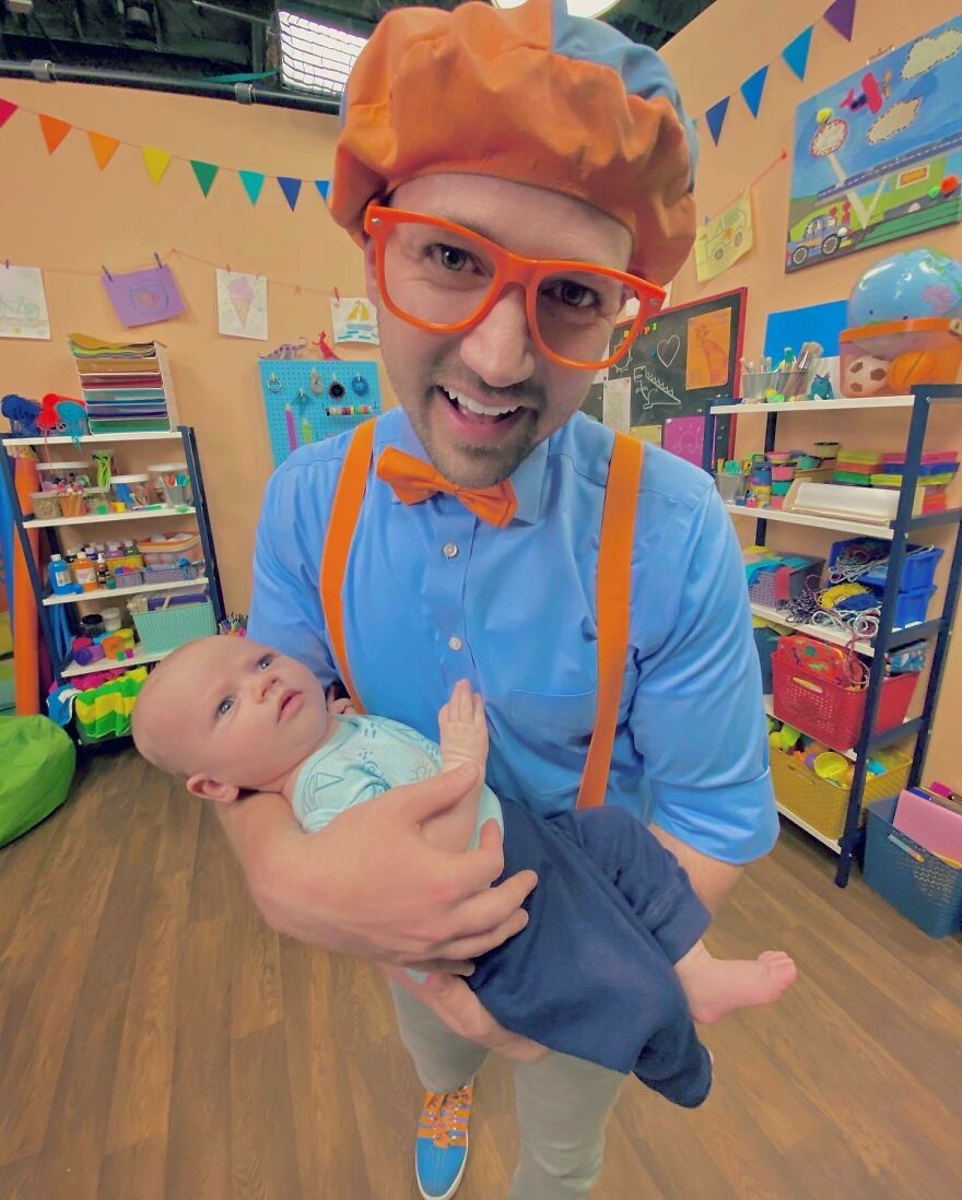 A Blippi wearing orange bow tie and glasses holding a baby in a colorful, playful room with educational decorations.