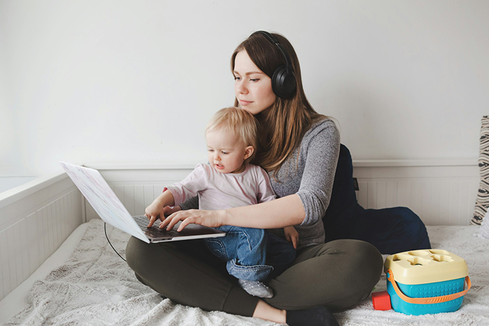 Woman working from home with child on lap, using laptop while wearing headphones in a casual home setting