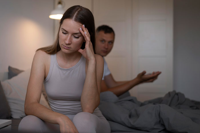 Woman looking upset sitting on the bed while boyfriend gestures behind her, relating to buying expensive gifts and Christmas conflict