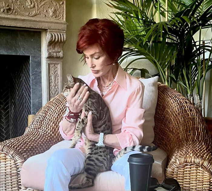 Sharon Osbourne sitting with a cat on her lap in a wicker chair next to indoor plants and a fireplace.