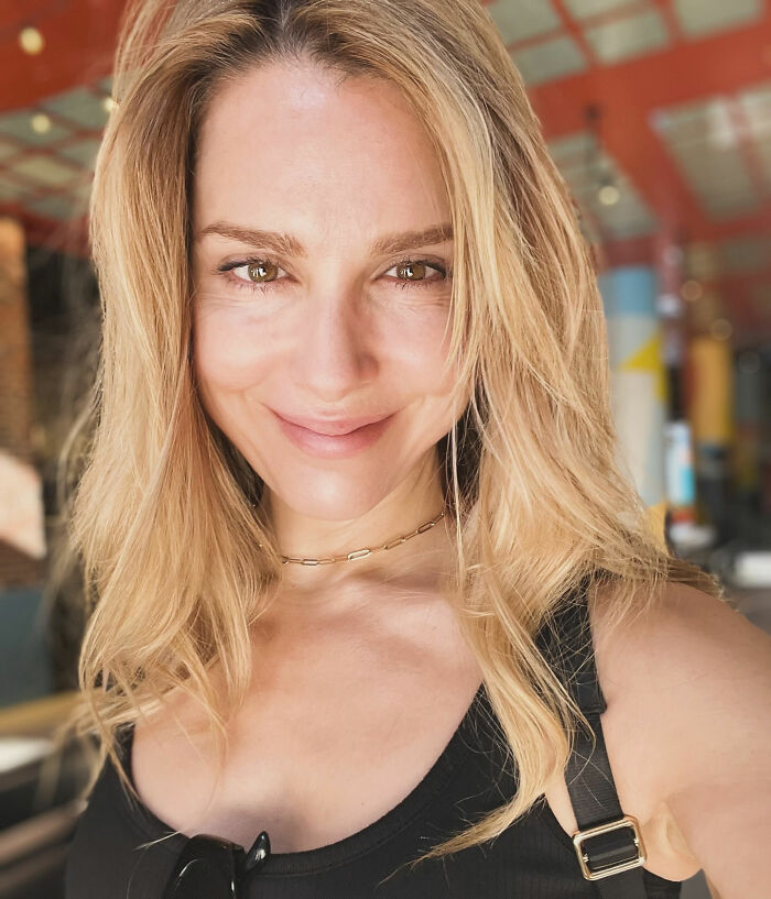 Woman with blonde hair and a black top smiling indoors, related to Stranger Things star and bullying involving Millie Bobby Brown.