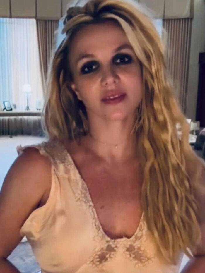Britney Spears with heavy eye makeup and long wavy hair in a beige top, looking different indoors.
