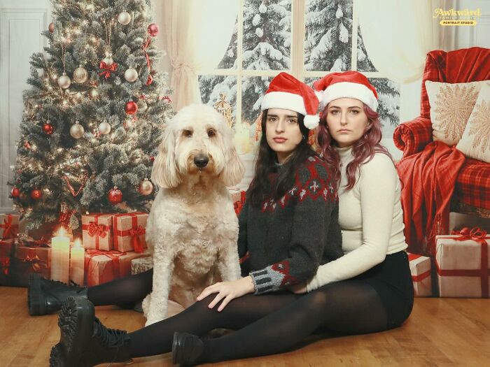 Two women wearing Santa hats and a dog posing awkwardly in a holiday-themed studio photo with Christmas decorations.