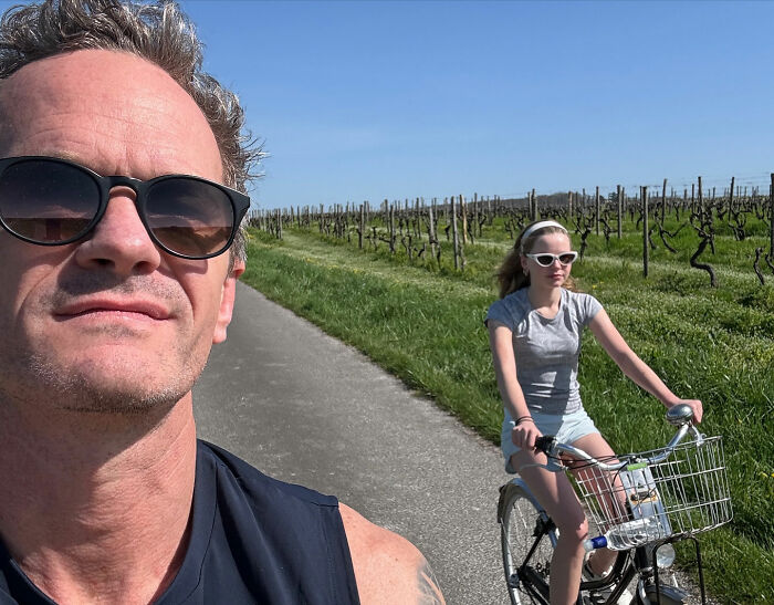 Neil Patrick Harris' daughter Harper, 15, riding a bike outdoors wearing sunglasses and a grey t-shirt.