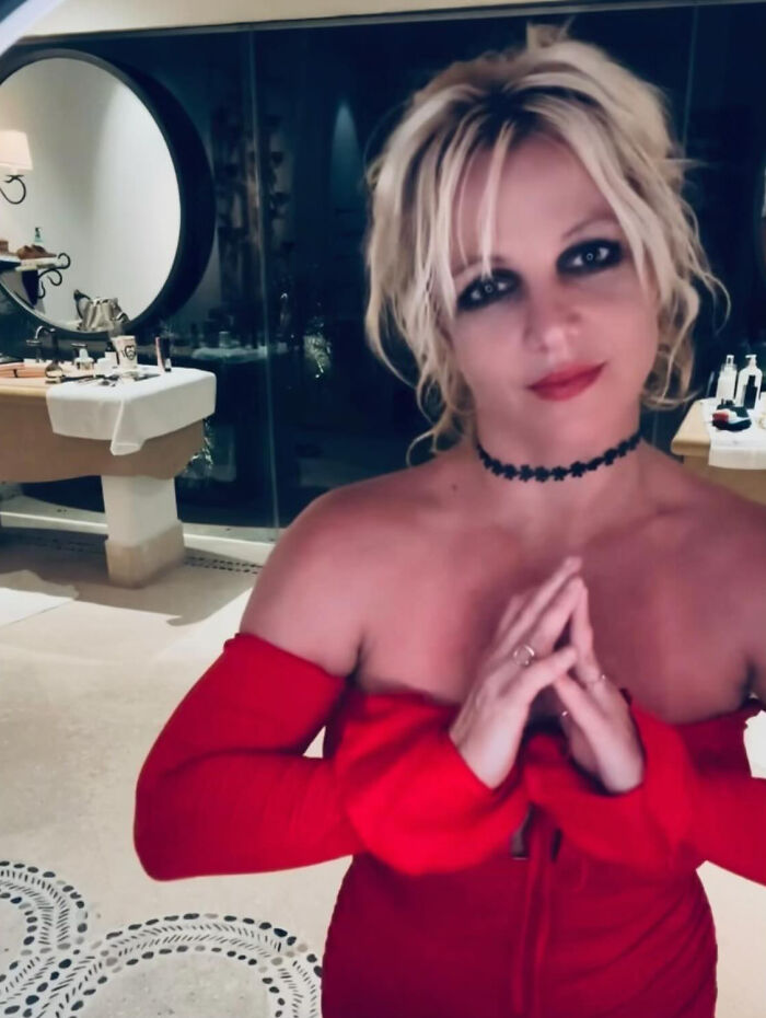 Britney Spears looking unrecognizable in a red dress with dark eye makeup, posing indoors with hands clasped near her chest.