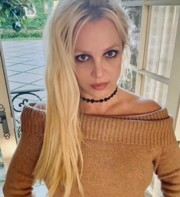 Britney Spears with long blonde hair and smoky eye makeup wearing a brown off-shoulder sweater indoors.