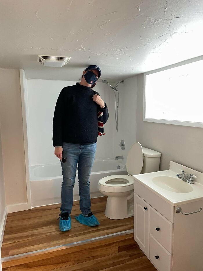 Person with face mask and shoe covers standing in a small bathroom of a Zillow home with low ceiling and wooden floor.