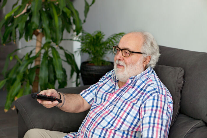 Elderly man in a plaid shirt sitting on a sofa holding a remote, representing infuriating examples of work habits.