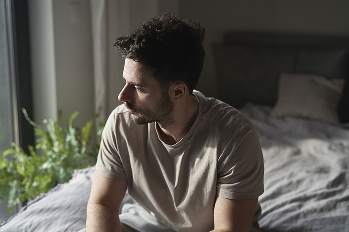 Man sitting on bed looking away thoughtfully, reflecting on girlfriend kissing another woman in front of friends.