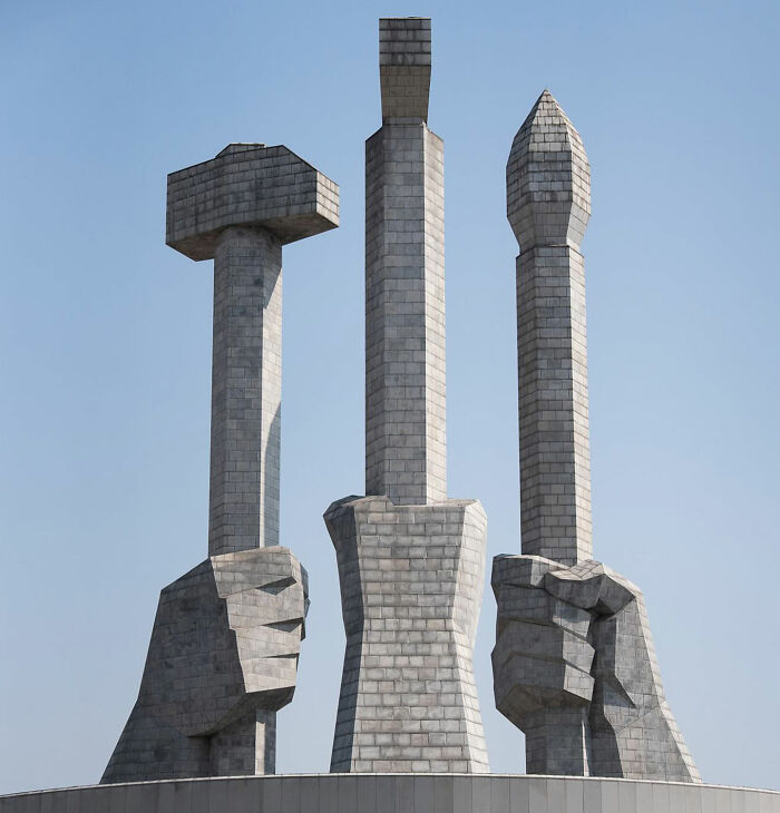 Monument in North Korea with three large hands holding a hammer, sickle, and brush under a clear blue sky.