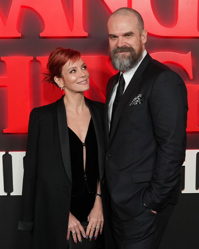 David Harbour and Stranger Things co-star smiling at event with red Stranger Things backdrop behind them.