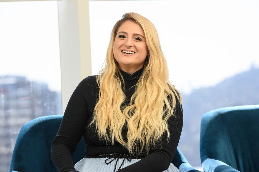 Meghan Trainor smiling during an interview, showcasing her long blonde hair and black top in a modern indoor setting.