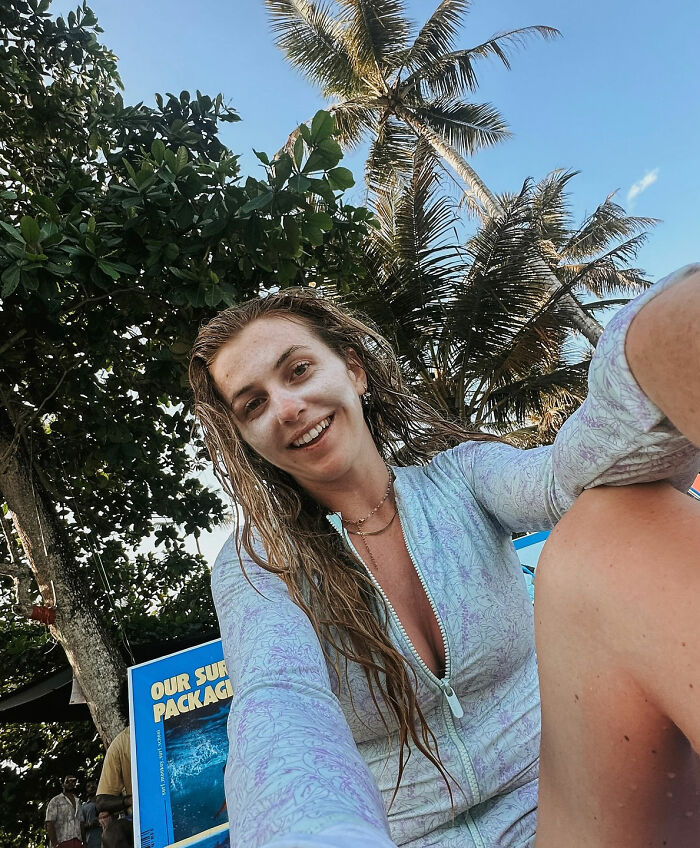 Woman solo traveling outdoors with palm trees in background, smiling and taking a selfie in casual beachwear.