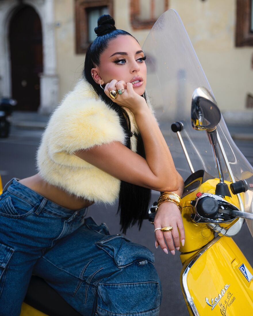 Camilla Araujo posing on a yellow scooter wearing a fluffy top and jeans, showcasing her net worth lifestyle.