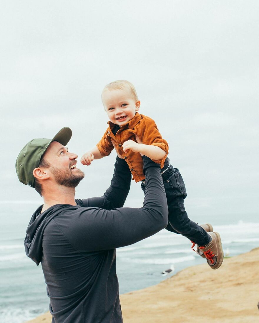 Stevin John wearing a cap lifting a smiling child by the ocean, illustrating the warmth behind how many Blippis actors have worn the bow tie.
