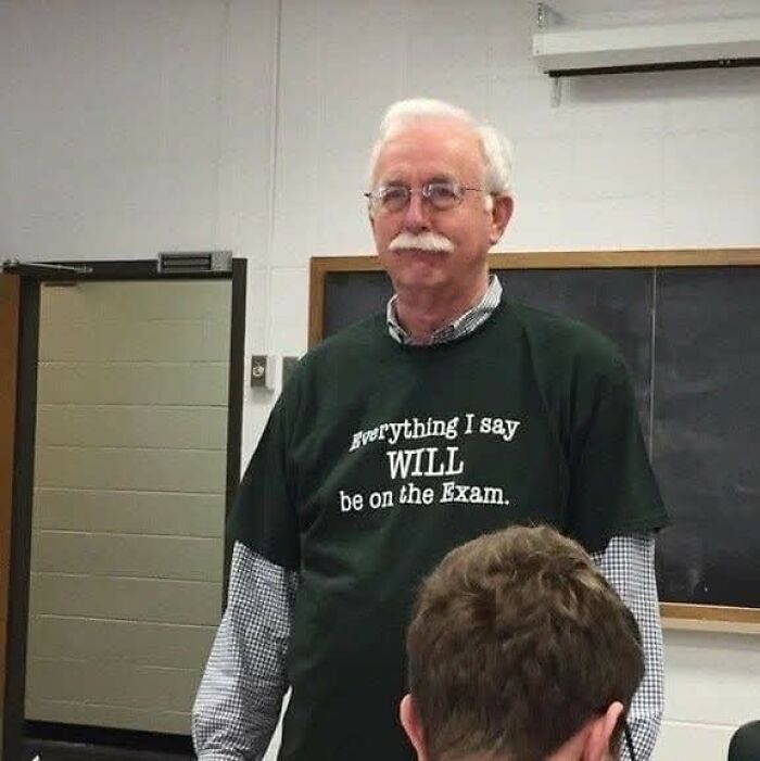 Older male teacher wearing glasses and a shirt with funny text, standing in classroom among hilarious educational memes.