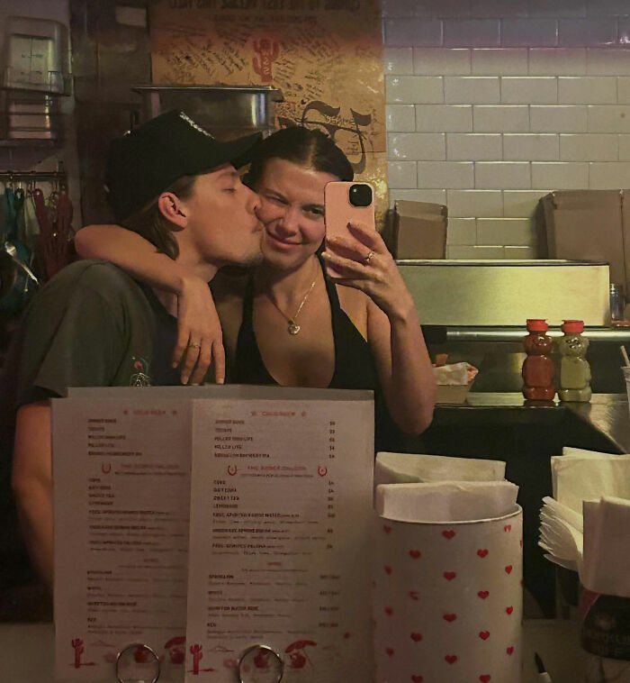 Millie Bobby Brown taking a selfie with a man kissing her cheek in a casual restaurant setting. Millie Bobby Brown taking a selfie with a man kissing her cheek in a casual restaurant setting.