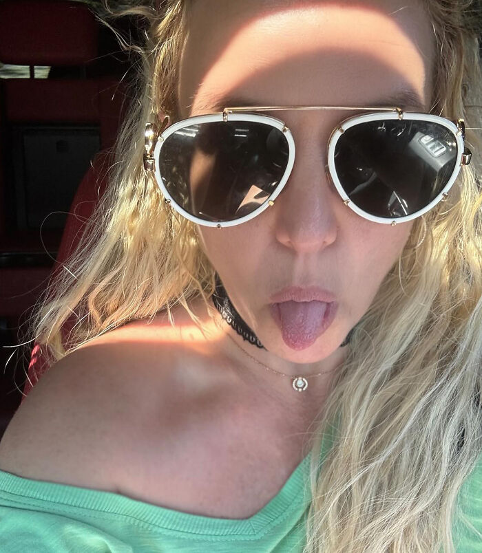 Britney Spears wearing large sunglasses and a green top, sticking out her tongue in a close-up selfie.